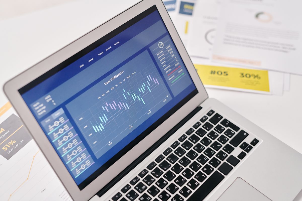 A laptop displaying financial charts beside printed reports